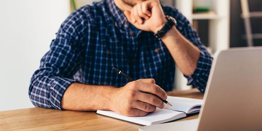 Man in a plaid shirt sitting at a desk, holding a pen and writing in a notepad while looking thoughtful. A laptop is open in front of him.